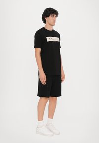 Svart T-skjorte med et beige logopanel med tekst, sammen med svarte shorts; modellen har på seg hvite sneakers og sokker, og står mot en hvit bakgrunn.