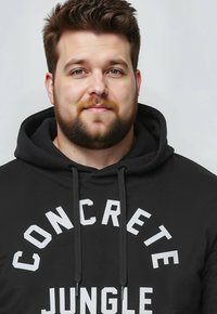 Sudadera negra con texto blanco "CONCRETE JUNGLE." Cuenta con capucha con cordón y una textura suave, diseñada para un ajuste casual.