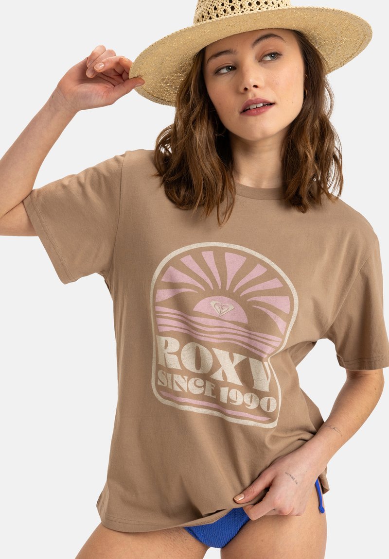 Hnědé bavlněné tričko s růžovým a bílým grafickým designem s motivem slunce a textem "ROXY SINCE 1990". Nošeno s modrými kalhotami.