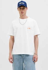 Witte katoenen t-shirt met korte mouwen en een zonopgangsafbeelding in oranje op de linkerkant van de borst. Gecombineerd met lichtblauwe denim shorts.