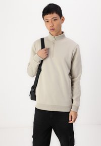 Beige mikina na zip s dlouhými rukávy, manžetami a lemem s žebrovaným vzorem, nošená přes tmavé cargo kalhoty, doplněná černou kabelkou přes rameno.