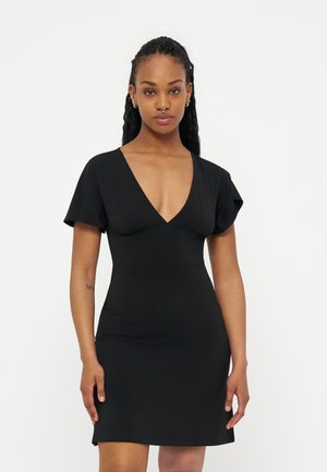 Ženska z dolgo pletenico, oblečena v kratko črno obleko z globokim V-izrezom in kratkimi cvetličnimi rokavi, stoji pred enostavnim ozadjem.