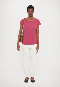 Fuchsia T-Shirt mit kurzen Ärmeln, kombiniert mit lockeren weißen Hosen, getragen zu flachen schwarzen Schuhen und einer braunen Umhängetasche. Einfaches, lässiges Outfit.