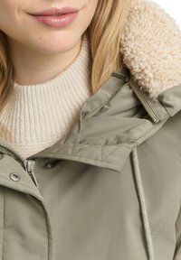 Grüne gesteppte Jacke mit Reißverschluss, ausgestattet mit einer cremefarbenen, mit Fleece gefütterten Kapuze, verstellbarem Kordelzug und einem beigefarbenen, gerippten Pullover darunter.