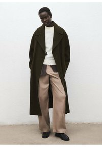 Manteau en laine vert foncé surdimensionné avec de grands revers, porté sur un pull léger et un pantalon beige large, complété par des chaussures noires.