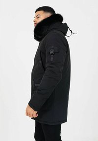 Svart parka-jacka med avtagbar huva foder av päls, sidofickor, texturerad finish och justerbar dragsnodd. Enkel design med funktionella detaljer.