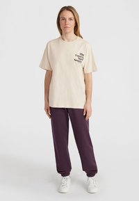 Beige katoenen t-shirt met zwarte tekst "The Future Surf Society!" op de borst, gecombineerd met ontspannen donkerpaarse sweatpants en witte sneakers.
