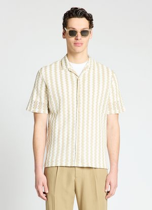 Mann trägt kurzärmliges beige-weiß gestreiftes Hemd, beige Hose und Sonnenbrille vor weißem Hintergrund.