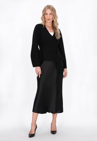 Top negru cu maneci lungi si evazate, cu un cordon la talie, asortat cu o fusta satinata neagra, lucioasa, pana la genunchi si pantofi cu toc ascutit, negri.