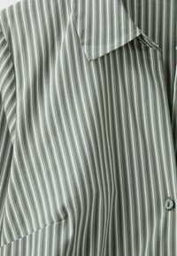 Gros plan d'une chemise grise à rayures verticales blanches, montrant le col, les boutons et une partie d'une poche.