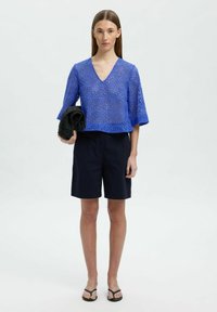 Blå, kortärmad, virkad topp med V-ringning och öppet mönster, ihop med mörka shorts och håller en svart stickad accessoar.