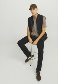 Black sleeveless vest over a gray shirt, paired with black pants and brown sneakers. A brown cap completes the look. Minimalist design.