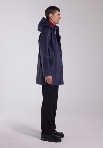 Man wearing navy raincoat with hood, red turtleneck, black pants, and black platform shoes, standing in side profile against plain background.