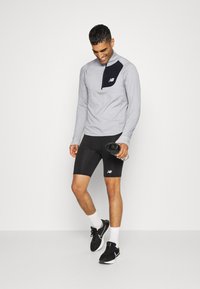 Camisola atlética de mangas longas cinza com um bolso preto no peito, combinada com calções pretos e sapatilhas pretas, segurando uma garrafa de água.
