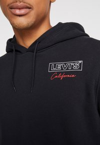 Sudadera negra de Levi's con un logo blanco y texto bordado en rojo que dice "California". Está hecha de una tela suave, tiene un corte relajado y capucha ajustable con cordón.