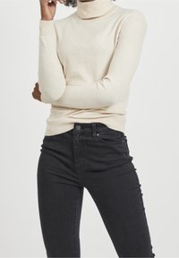 Maglione a dolcevita beige chiaro con maniche lunghe, polsini e orlo a coste. Abbinato a jeans neri aderenti con vita con bottoni e tasche.