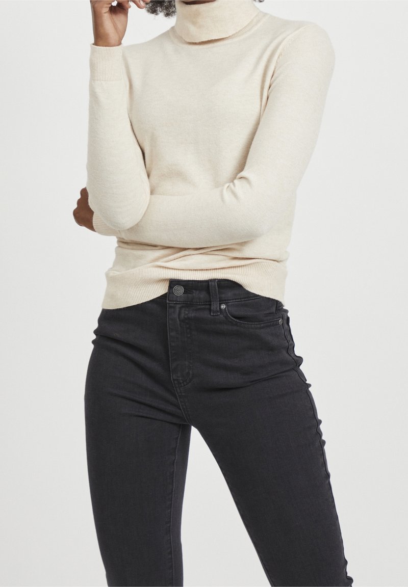 Maglione a dolcevita beige chiaro con maniche lunghe, polsini e orlo a coste. Abbinato a jeans neri aderenti con vita con bottoni e tasche.