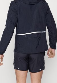Personne portant une veste légère noire et un short noir vue de dos sur un fond uni.