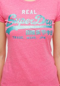 Camiseta rosa de tela suave con un diseño gráfico texturizado y brillante en turquesa que incluye "REAL SuperDry" y texto adicional abajo.