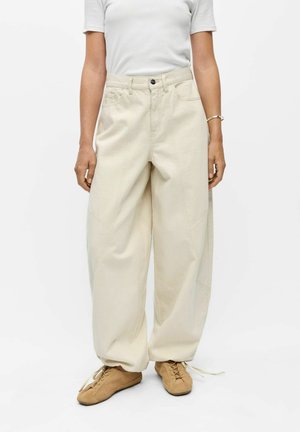 Personne portant un pantalon large ample blanc cassé, une chemise blanche à manches courtes, des chaussures lacées couleur beige et un bracelet argenté au poignet droit.