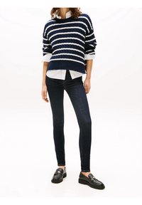 Mujer vestida con un suéter de rayas azul marino y blanco, camisa blanca, jeans ajustados oscuros y mocasines de cuero negro, de pie contra un fondo blanco.
