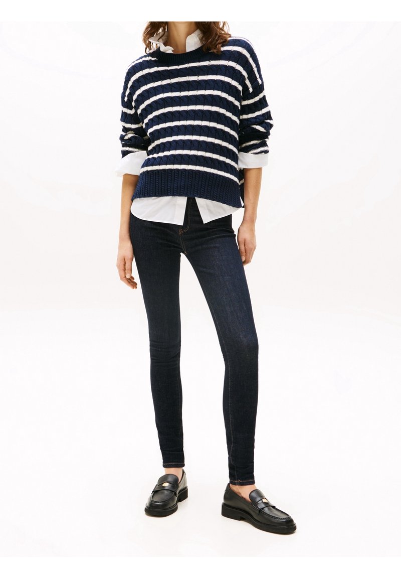 Mujer vestida con un suéter de rayas azul marino y blanco, camisa blanca, jeans ajustados oscuros y mocasines de cuero negro, de pie contra un fondo blanco.