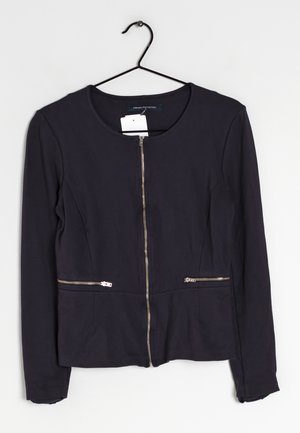 Schwarze, taillierte Jacke mit rundem Ausschnitt, langen Ärmeln und einem zentralen Reißverschluss. Verfügt über zwei mit Reißverschluss gesicherte Taschen auf Taillenhöhe. Glatte Stofftextur.