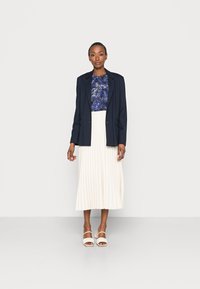 Blazer azul-marinho sobre uma blusa com padrão azul, combinado com uma saia midi plissada creme e sandálias de salto brancas. Texturas suaves e corte ajustado.
