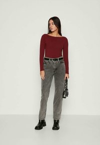 Burgundy long-sleeve top paired with gray high-waisted jeans, black belt, and black ankle boots. Metallic accents on bag. Simple background.