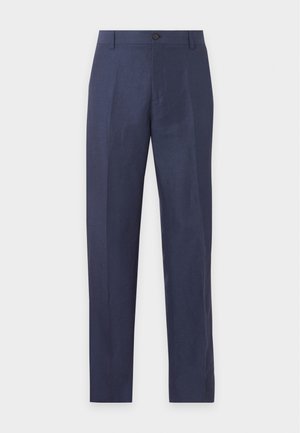 Homme debout, les mains dans les poches, portant un polo en maille blanc à manches courtes, un pantalon bleu marine et des mocassins marron foncé sur un fond uni.