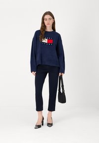 Maglione over-size blu navy con logo bianco e rosso, abbinato a pantaloni scuri e tacchi alti neri, mentre tiene una piccola borsa nera.