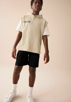 Dreng, der står og har på en beige ærmeløs hættetrøje med teksten "SUBSCRIBE", hvid kortærmet skjorte, sorte shorts, hvide sokker og hvide sneakers.