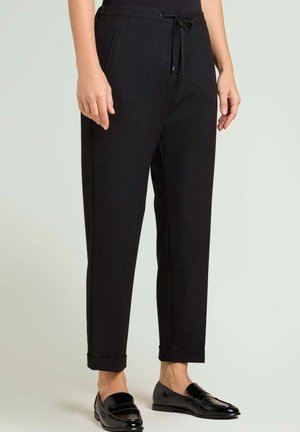 Pantalon noir fuselé avec une taille à cordon, des poches latérales et des revers retroussés, assorti à des mocassins noirs. Texture de tissu lisse.