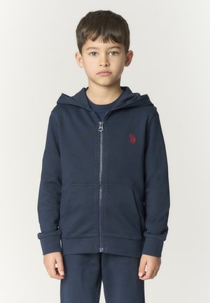 Ragazzo con capelli castani corti che indossa una felpa con cappuccio blu navy con cerniera e un piccolo logo rosso sul petto, in piedi davanti a uno sfondo chiaro e semplice.