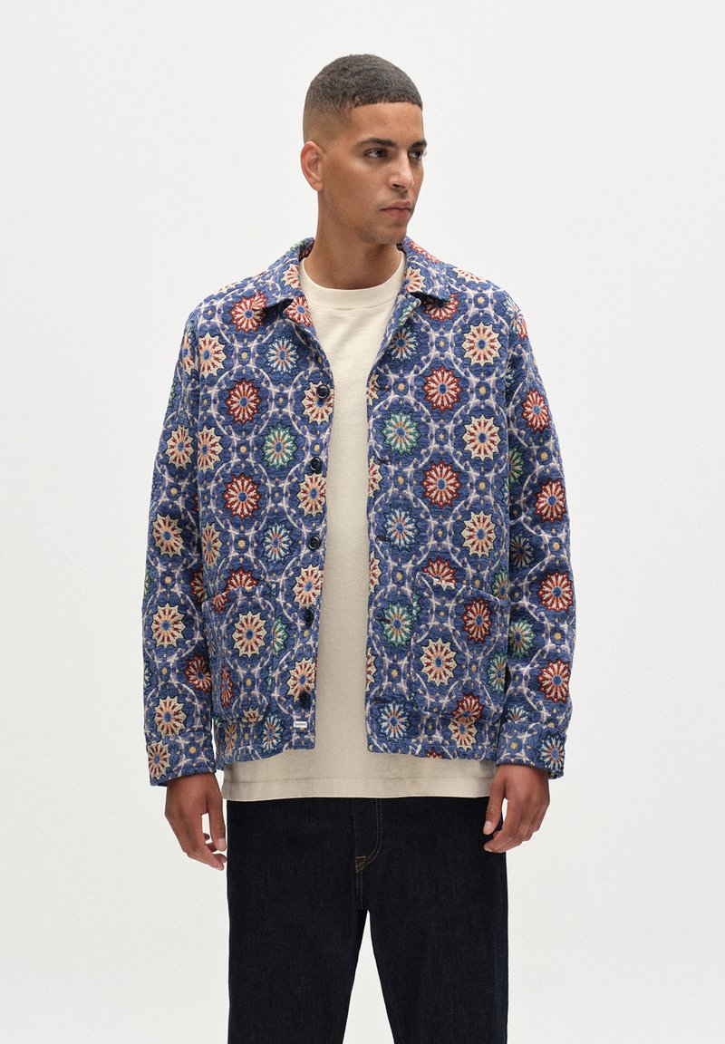 Musterjacke mit blauer Basis, die blumige Motive in Rot, Weiß, Grün und Orange zeigt. Mit Knopfleiste und zwei frontalen Taschen.