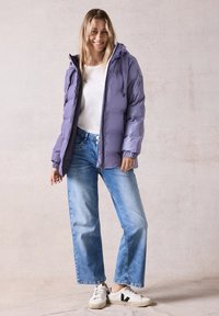 Doudoune violette claire avec une capuche, fermeture éclair et poignets élastiques, assortie à un jean bleu et des baskets blanches avec des accents noirs.