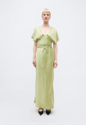 Naine seisab, kandes helerohelist satiinist maksikleitpuhvis varrukate, sügava V-kaeluse, vööga piht salvestatud ja mustade teravate jalanõudega.