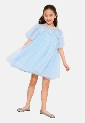 Pige, der smiler og snurrer rundt i en lyseblå kjole med pufærmer, blomstrede blonde detaljer og glitrende sølvfarvede flade sko på en hvid baggrund.