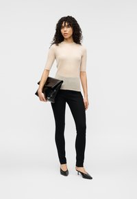 Beiges, geripptes Strickoberteil mit kurzen Ärmeln, kombiniert mit schwarzen, schlanken Hosen und schwarzen, spitzen High Heels. Hält eine schwarze Clutch.