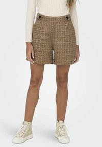 Shorts en tweed marron avec un motif texturé, taille haute, et deux boutons noirs proéminents. Associés à un haut côtelé beige clair et des baskets beiges.