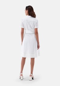 Weißes Hemdkleid mit kurzen Ärmeln, Spitzen-Augenlochdetails, tailliertem Schnitt und weit ausgestellt Rock, mit einem Saumverzierung und einer glatten Textur.