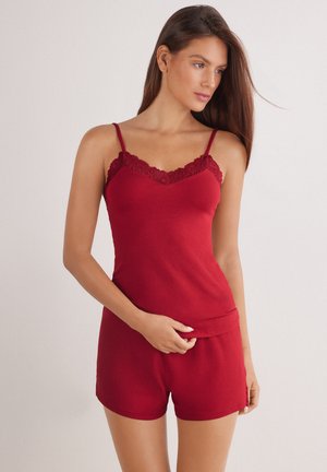 Kvinde iført en rød camisole med blonder og matchende shorts, stående med den ene hånd hvilende på hendes talje og kiggende til siden.