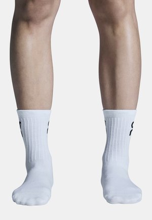 Vitaid atletiska strumpor med ribbad textur, med svart varumärke på sidan, vilande på bara fötter mot en ljus bakgrund.