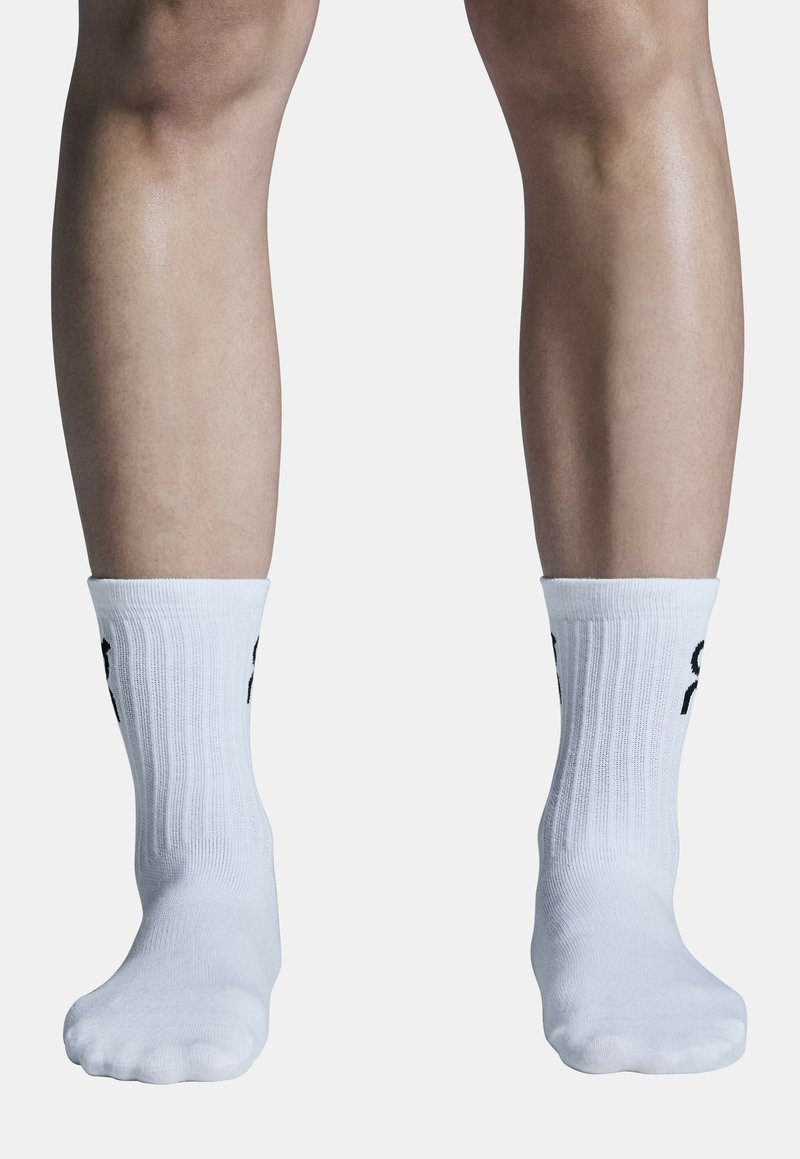 Hvide sportsstrømper med ribbet tekstur, prydet med sort branding på siden, hvilende på bare fødder mod en lys baggrund.