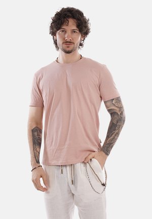 Uomo con capelli ricci che indossa una maglietta rosa chiaro e pantaloni bianchi, mostra tatuaggi dettagliati su entrambe le braccia, in piedi contro uno sfondo bianco.