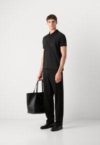 Μαύρο πόλο μπλουζάκι με τσέπη μπροστά και λογότυπο, σε συνδυασμό με μαύρους φαρδιούς παντελόνες και γυαλιστερά μαύρα παπούτσια, κρατώντας μια μαύρη τσάντα tote.