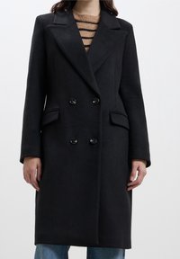 Cappotto nero doppiopetto realizzato in tessuto testurizzato, con rever a punta, due tasche frontali e bottoni a contrasto.