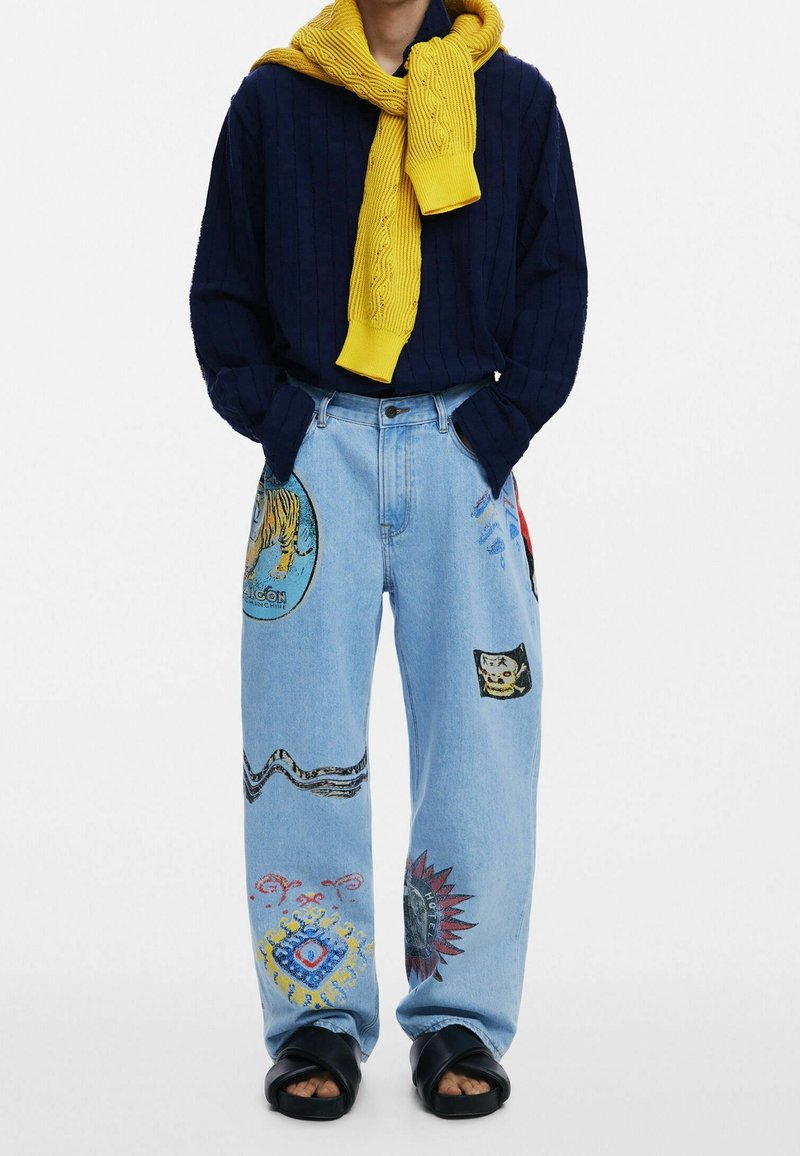 Personne portant un jean bleu clair avec des patchs colorés, un pull bleu marine avec un pull jaune drapé sur les épaules et des sandales noires.