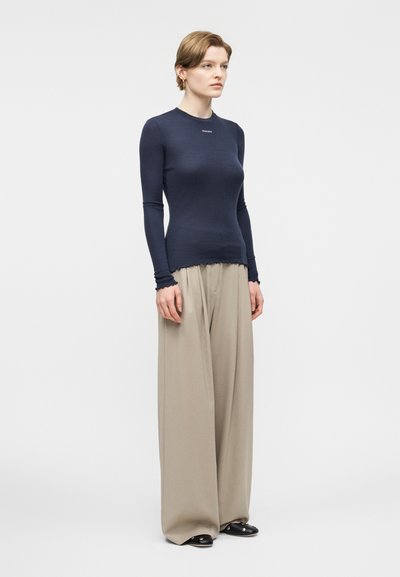 Γυναίκα που στέκεται, φορώντας μακρυμάνικη εφαρμοστή μπλούζα σε ναυτικό μπλε χρώμα και φαρδιά μπεζ παντελόνια με μαύρα παπούτσια, σε απλό φωτεινό φόντο.