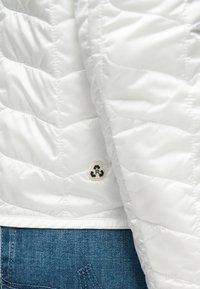 Λευκό καπιτονέ μπουφάν με λογότυπο από ανακυκλωμένο υλικό κοντά στο τελείωμα, που καλύπτει μερικώς μπλε τζιν τζιν.